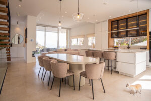 Comedor y cocina premium con isla blanca, asientos beige y iluminación moderna; espacio luminoso con vistas y un perro. INKO Cocinas