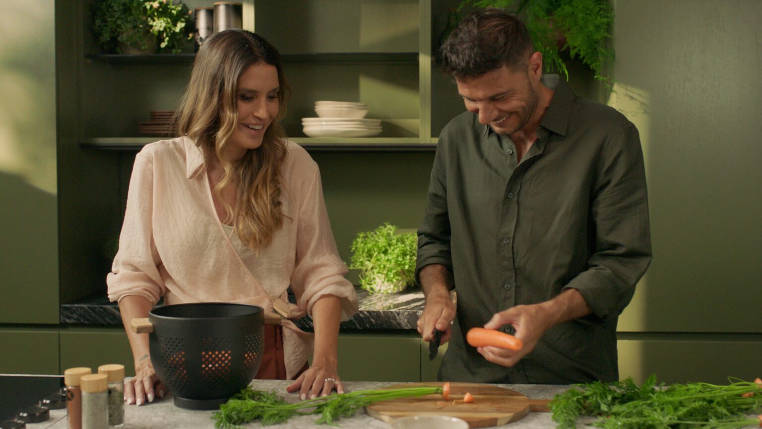 INKO Cocinas pareja en una cocina de diseño premium con muebles verde-oliva, preparando verduras en una tabla junto a una olla negra