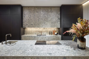 Cocina de diseño sofisticado y calidad premium con encimeras y frontal en piedra gris, muebles oscuros e iluminación cálida. INKO Cocinas
