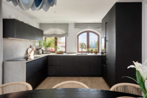 INKO | Fabricantes de Cocinas en Málaga y Diseño a Medida | Cocina contemporánea en negro mate con encimeras claras, fregadero con grifo negro bajo una gran ventana arqueada; diseño premium INKO Cocinas